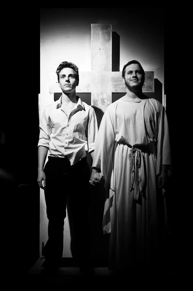 Black and white photo of two people standing in front of a cross-shaped statue, one person in casual clothing and the other in a religious robe, holding hands.