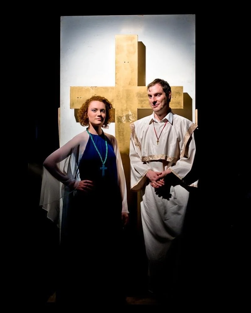 A man and woman in religious costumes standing in front of a large gold cross on a white background.