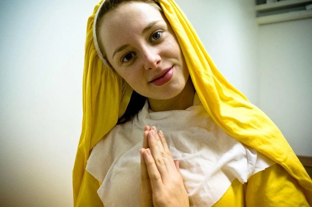 A woman dressed as Jesus Christ with a yellow robe and head covering, holding her hands in prayer.