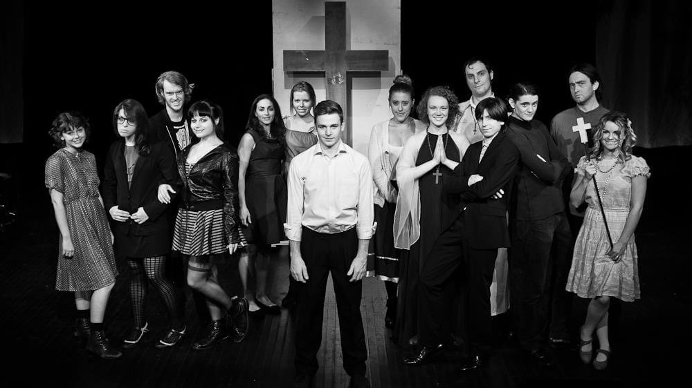 Group of people on stage with a cross in the background, some dressed as nuns and others in casual or gothic attire, in black and white.