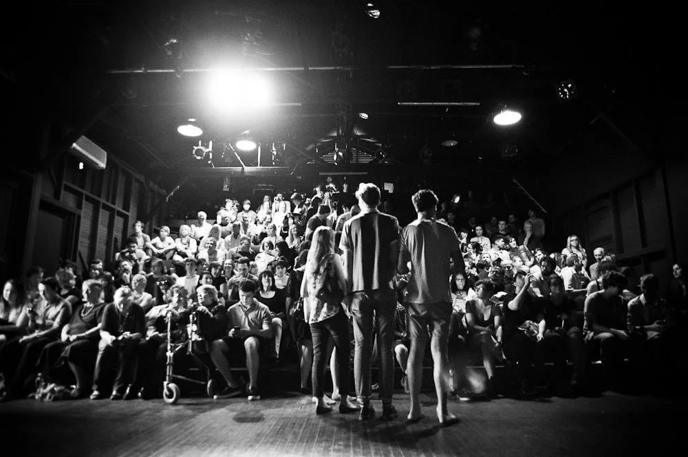 Black and white photo of a theater or comedy club filled with audience members seated and watching a performance. Three performers stand on stage facing the audience, backlit by bright lights.
