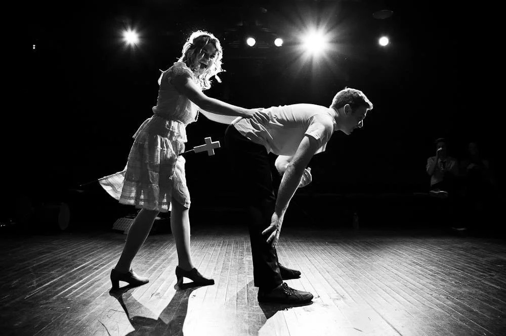 Girl pulling boy on a stage during a performance with spotlight and audience members in background.