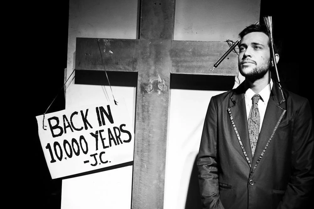 A man in a suit stands next to a wooden cross with a sign that says "Back in 10,000 years - JC."