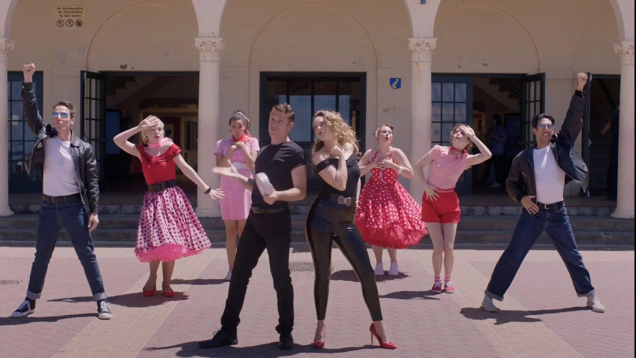 A group of performers in vintage 1950s style clothing dancing outside a building with white columns and arches.