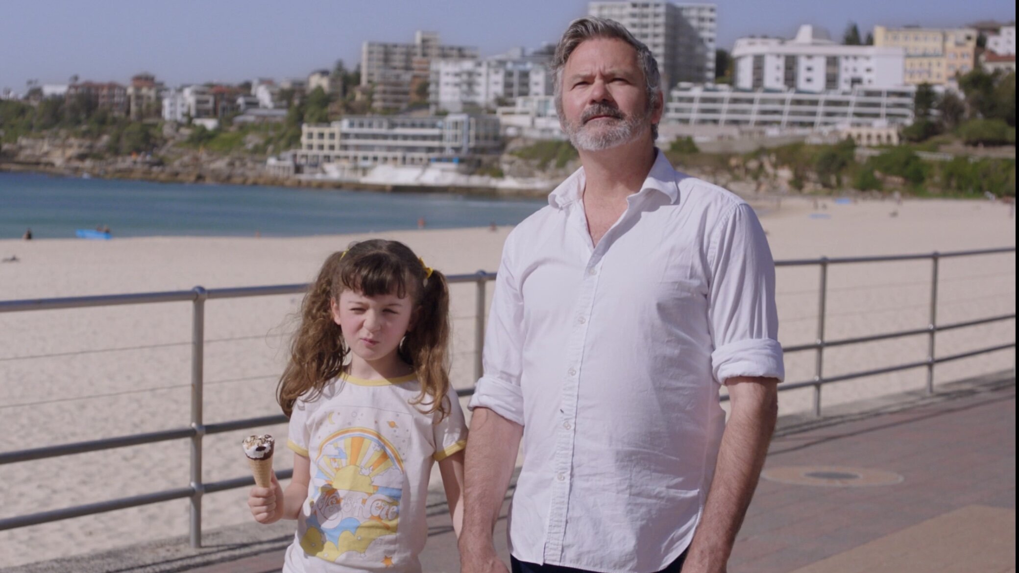 A man and a young girl walking along a beach boardwalk with buildings in the background.