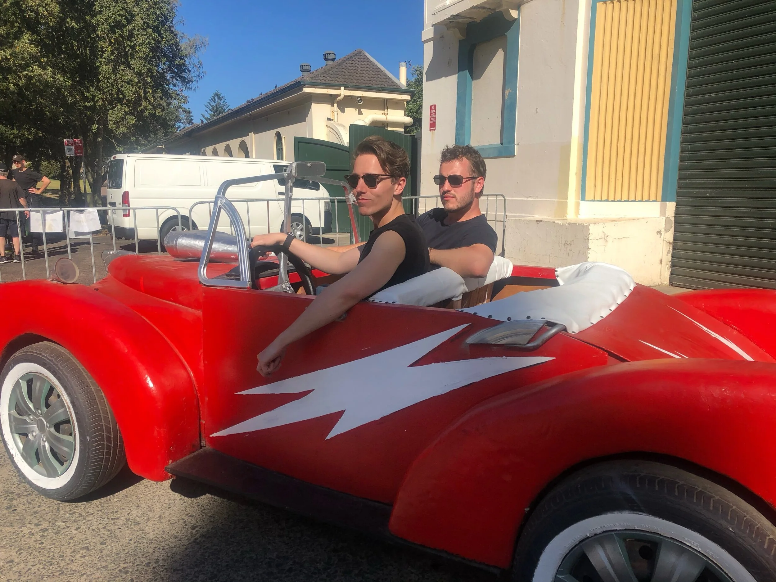 Two men sitting in a red vintage car with white detailing parked on a street, with trees, a white van, and a building with a sign in the background.
