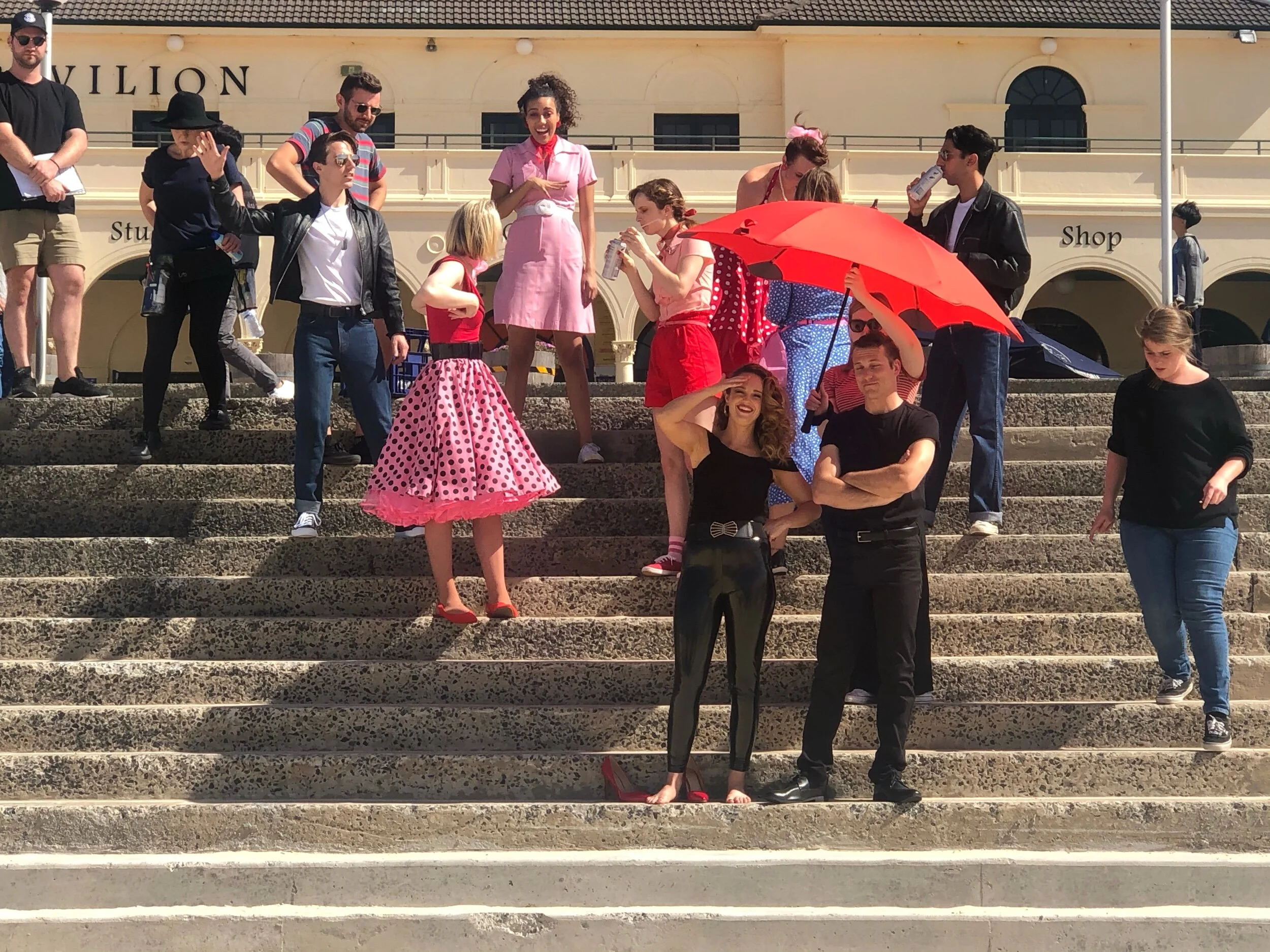 A group of people on outdoor steps, some dressed in retro or pin-up style clothing, including polka dot skirts and vintage hair accessories, with one woman holding a red umbrella. The background shows a building with the partially visible sign readin