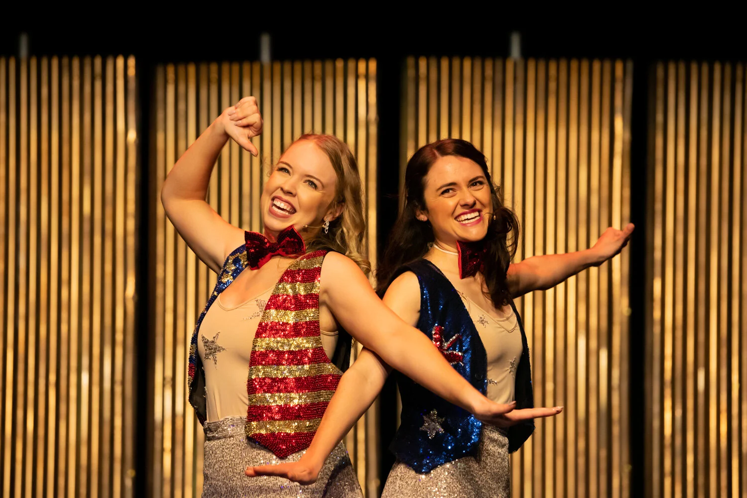 Two women are performing on stage, dressed in sparkly costumes with star and stripe patterns, wearing red bow ties, smiling, with a gold and black striped background.