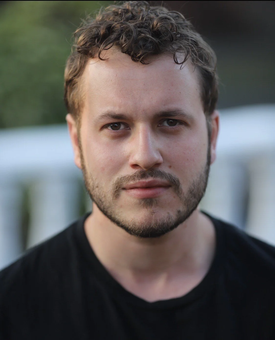 Close-up of a young man with curly brown hair and a beard, wearing a black shirt, outdoors with blurred greenery in the background.