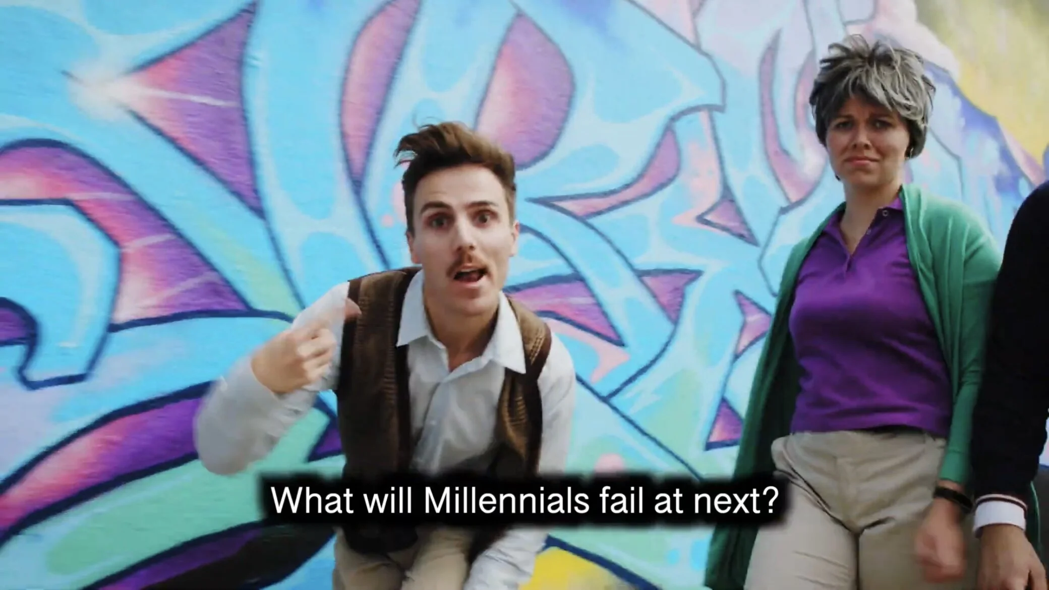 Two young adults, a man and a woman, standing in front of a colorful graffiti wall. The man has brown hair styled upwards, wears a white shirt, beige pants, and carries a backpack. The woman has short, wavy gray hair, wears a purple shirt with a gree