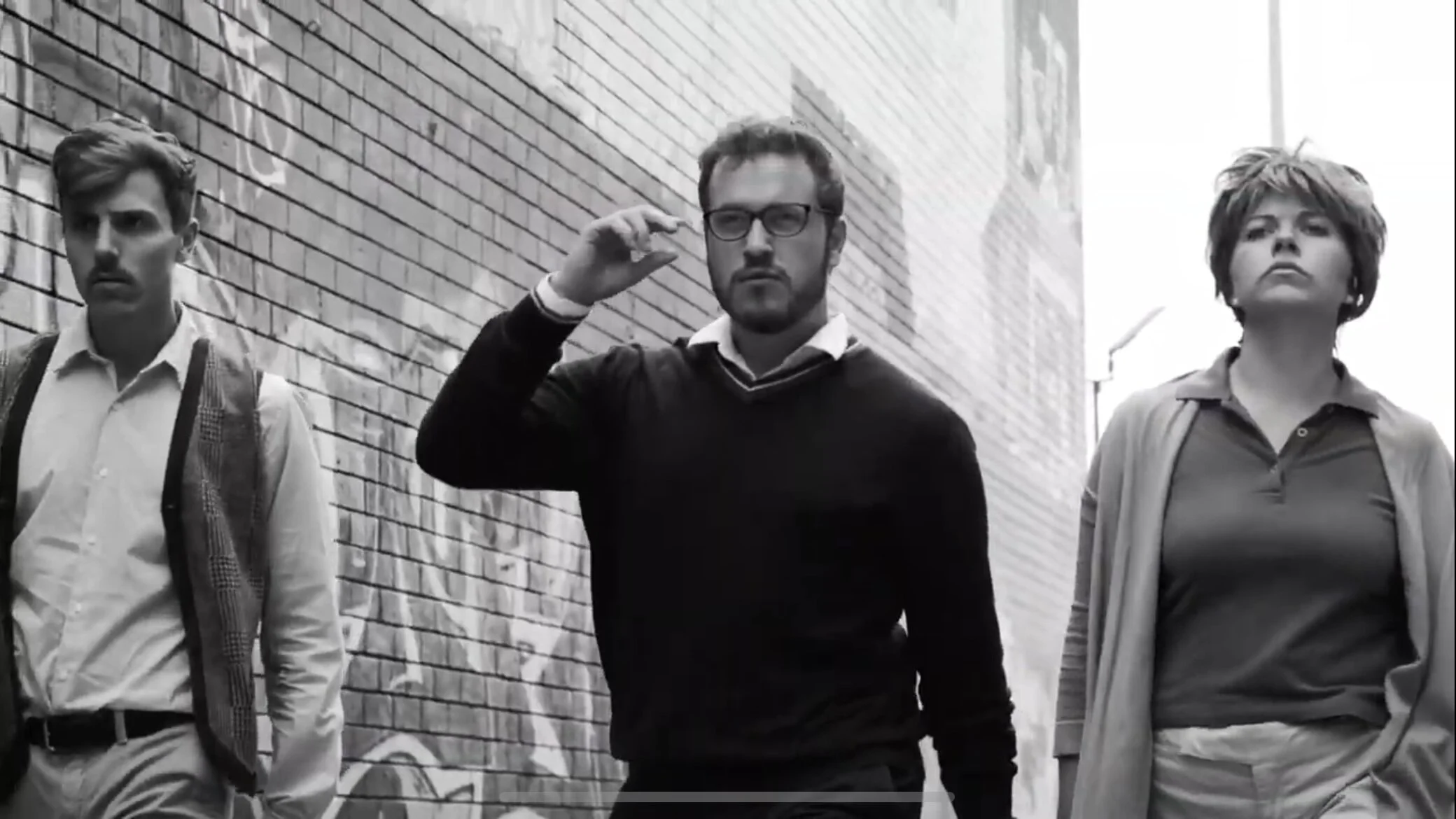 Three people walking along a brick wall, black and white photo. The person in the middle is adjusting glasses, while the others walk on either side.