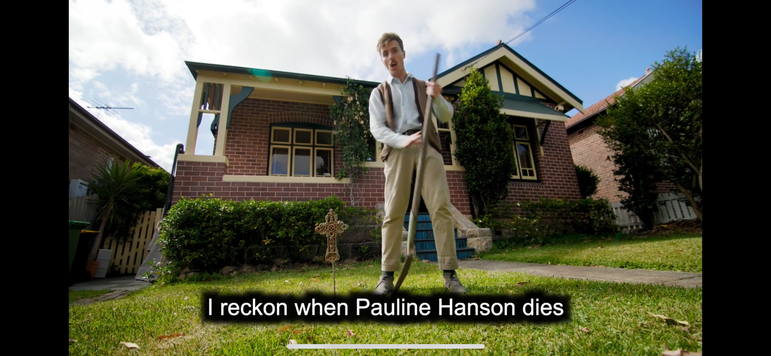 A young man with short hair, wearing a white shirt and beige pants, stands on a lawn holding a stick, in front of a brick house with a porch, greenery, and trees. Subtitles at the bottom read: 'I reckon when Pauline Hanson dies'.