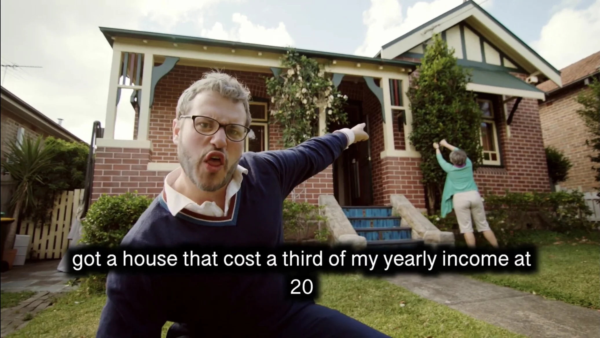 A man with glasses pointing at a house with a woman pruning a tree in the front yard.