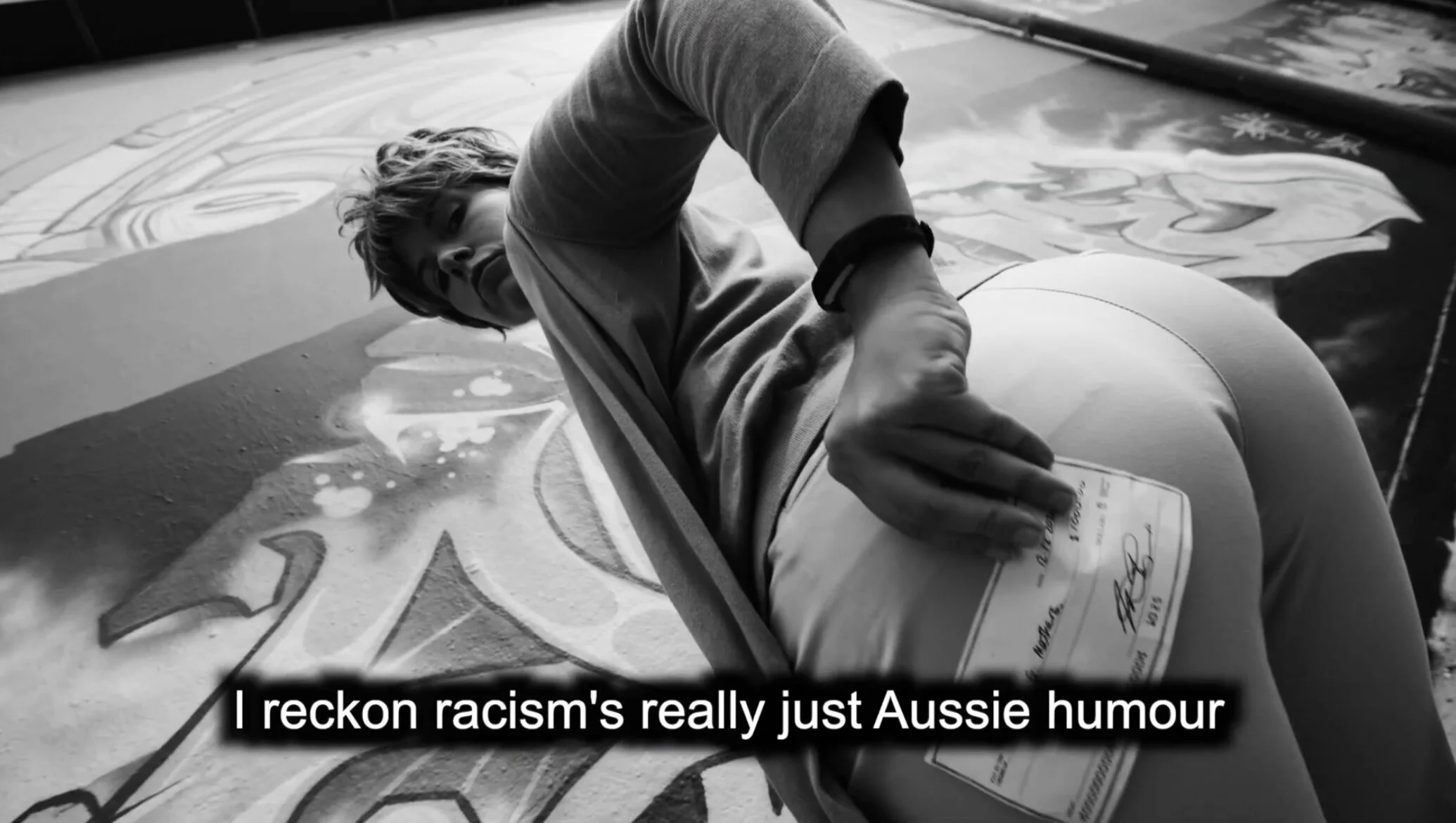 A person painting on a large surface decorated with colorful artwork, holding a container of paint, with subtitles that read 'I reckon racism's really just Aussie humour'.