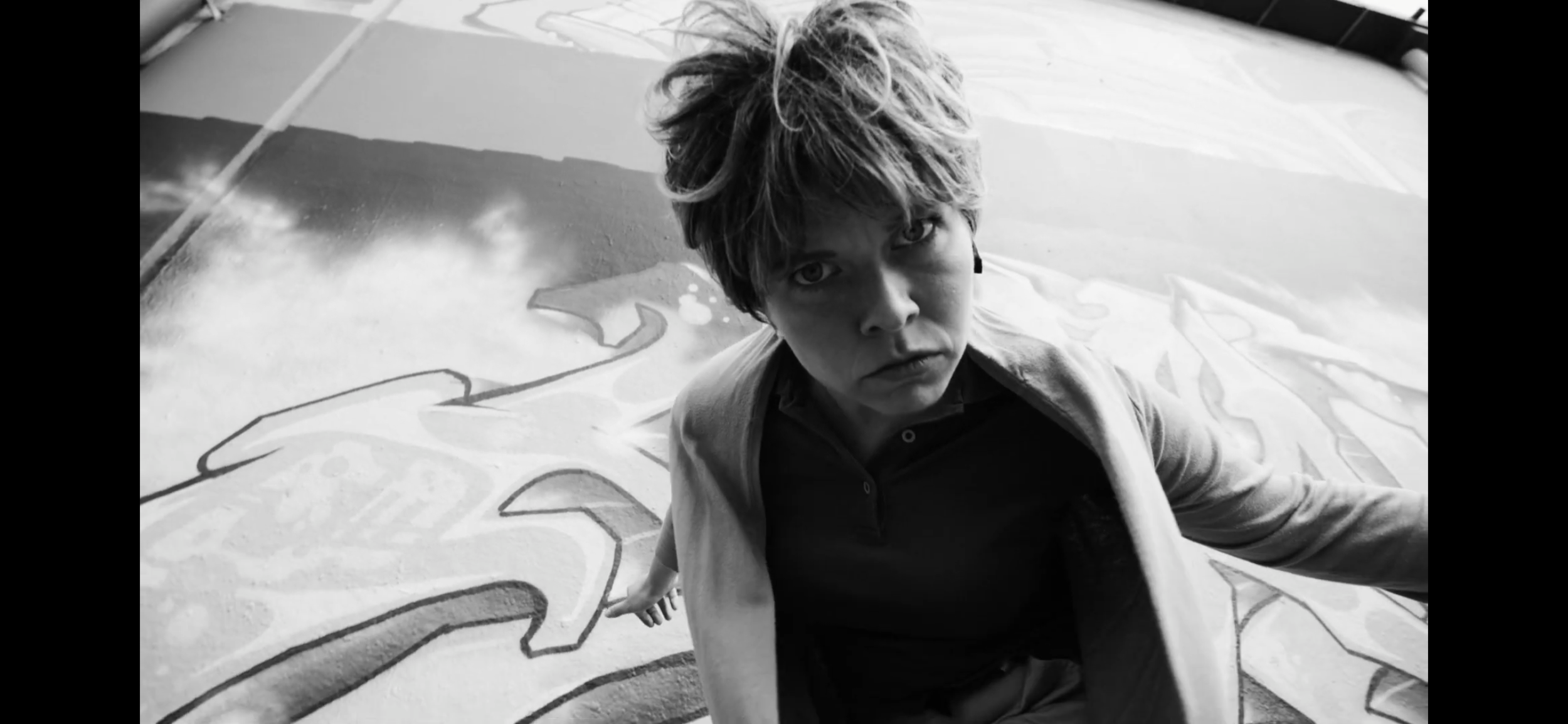 A woman with short, messy hair looking directly at the camera while sitting on the ground. There is a large piece of artwork or a mural with cartoonish characters and colorful markings behind her.
