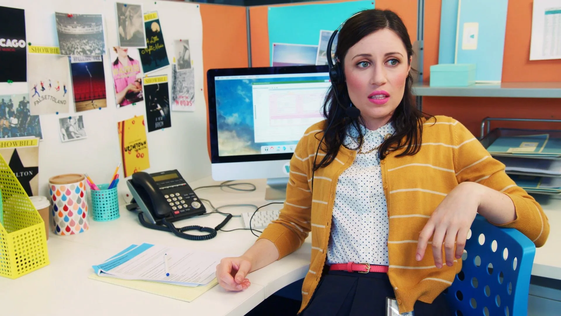 A woman with dark hair wearing a yellow striped cardigan and polka dot shirt, sitting at her desk in an office, wearing a headset, looking to her right with a surprised expression. Her desk has a black telephone, colorful pens in a holder, papers, and a computer monitor displaying a webpage. The background includes a bulletin board with photos and posters and office supplies.