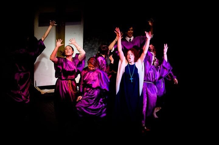 Group of women dressed in purple robes singing with their hands raised, a woman in the center with a cross necklace also singing with arms raised, on a dark stage.