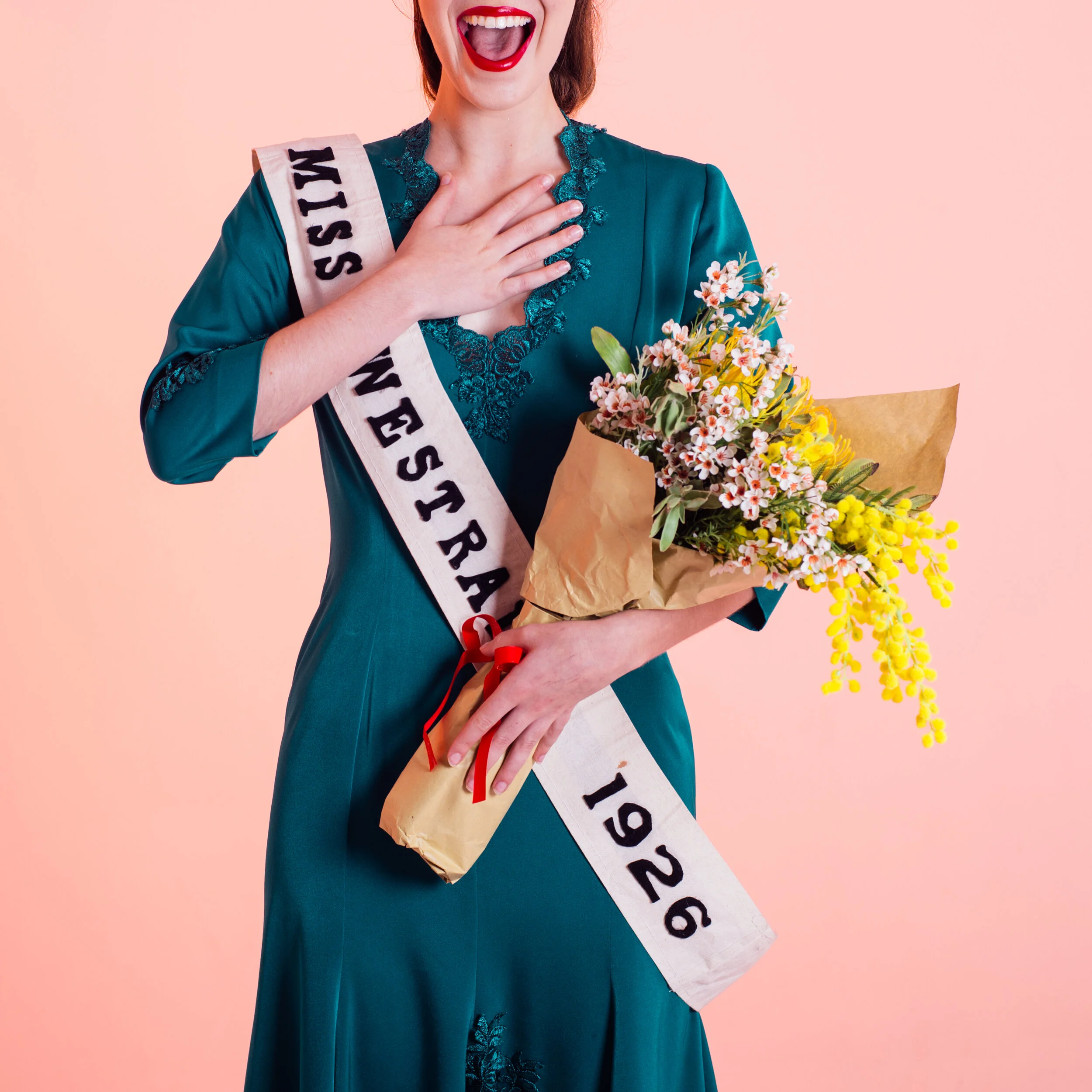 A woman with red hair, wearing bright red lipstick and a teal dress with lace accets, is smiling broadly with her mouth open, one hand over her chest, and holding a bouquet of white, yellow, and pink flowers wrapped in brown paper. She is also wearing a sash that reads 'MISS in black letters. The background is a solid light pink color.