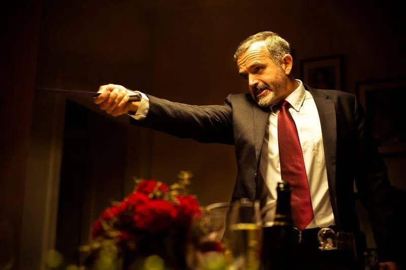 A man in a suit and red tie pointing a knife, with a serious expression, in a dimly lit room.