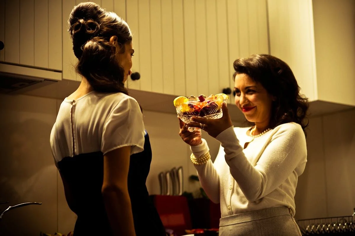 Two women in a kitchen, one in retro attire holding a colorful bowl of dessert, smiling at the other woman.