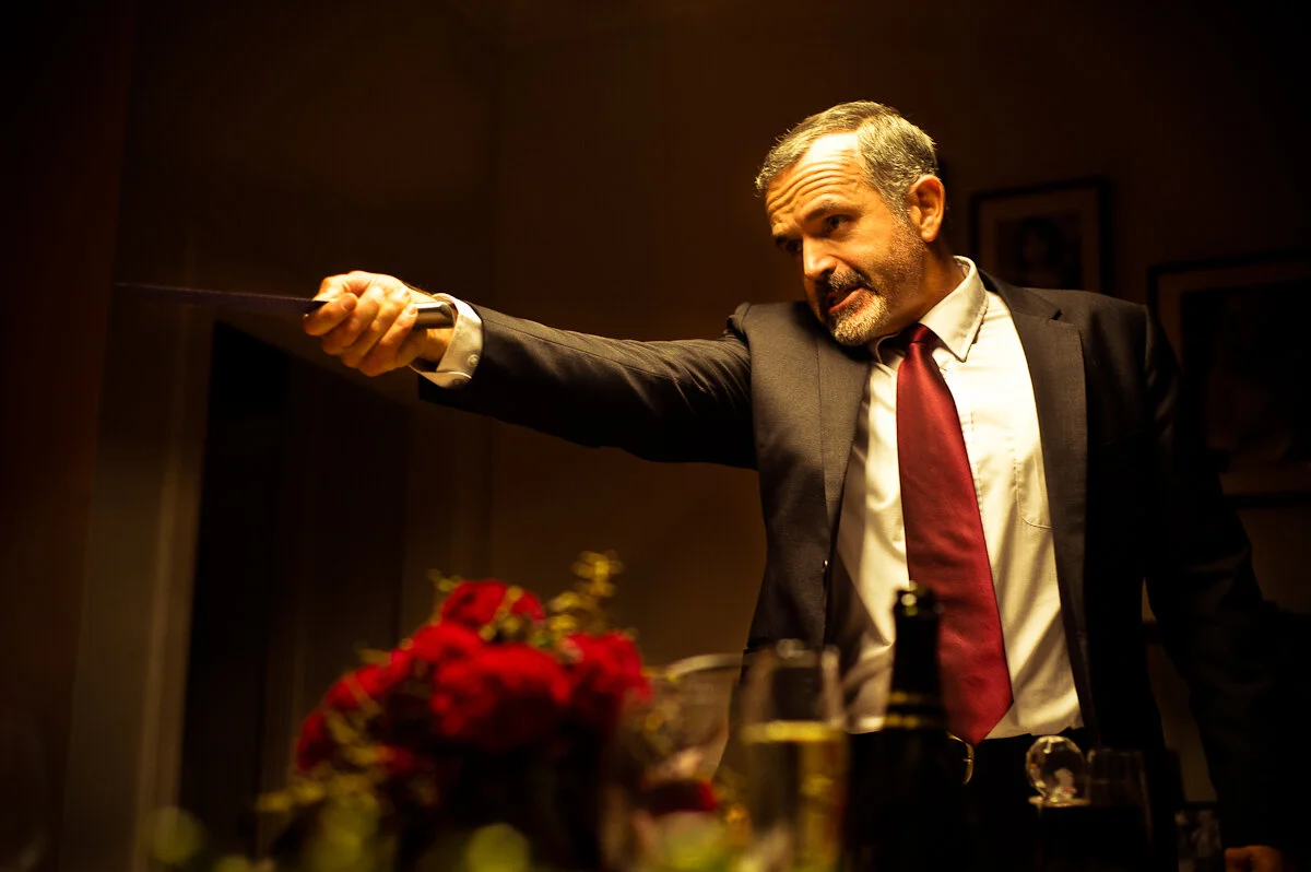 A man in a suit and red tie pointing a gun in a tense setting, with bottles and flowers on the table.