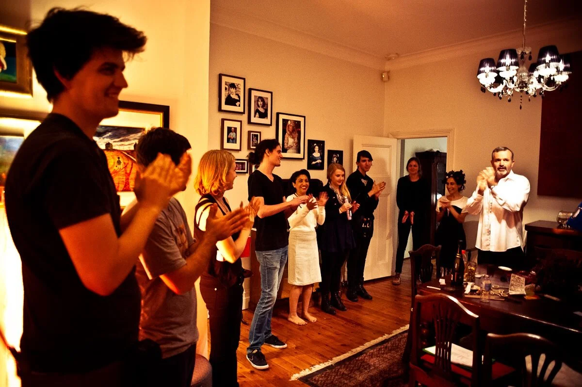 A group of people standing in a line in a cozy living room, clapping and smiling, with artworks on the wall and a chandelier hanging from the ceiling.