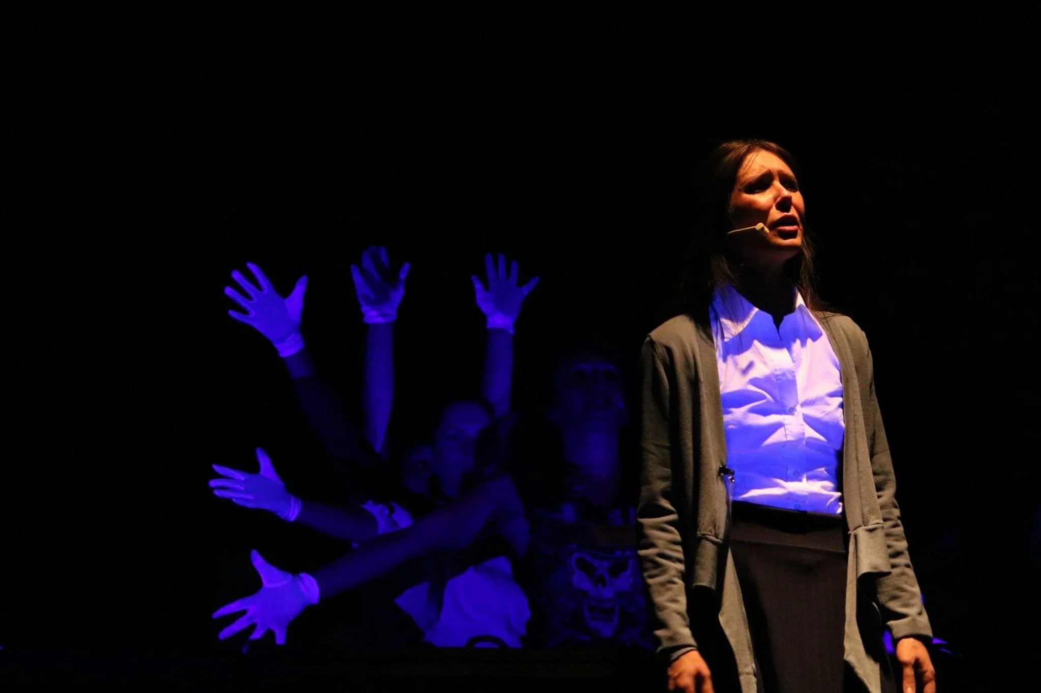 A woman standing on stage with a microphone, illuminated by stage lighting, with multiple hands wearing blue gloves raised in the background against a dark backdrop.