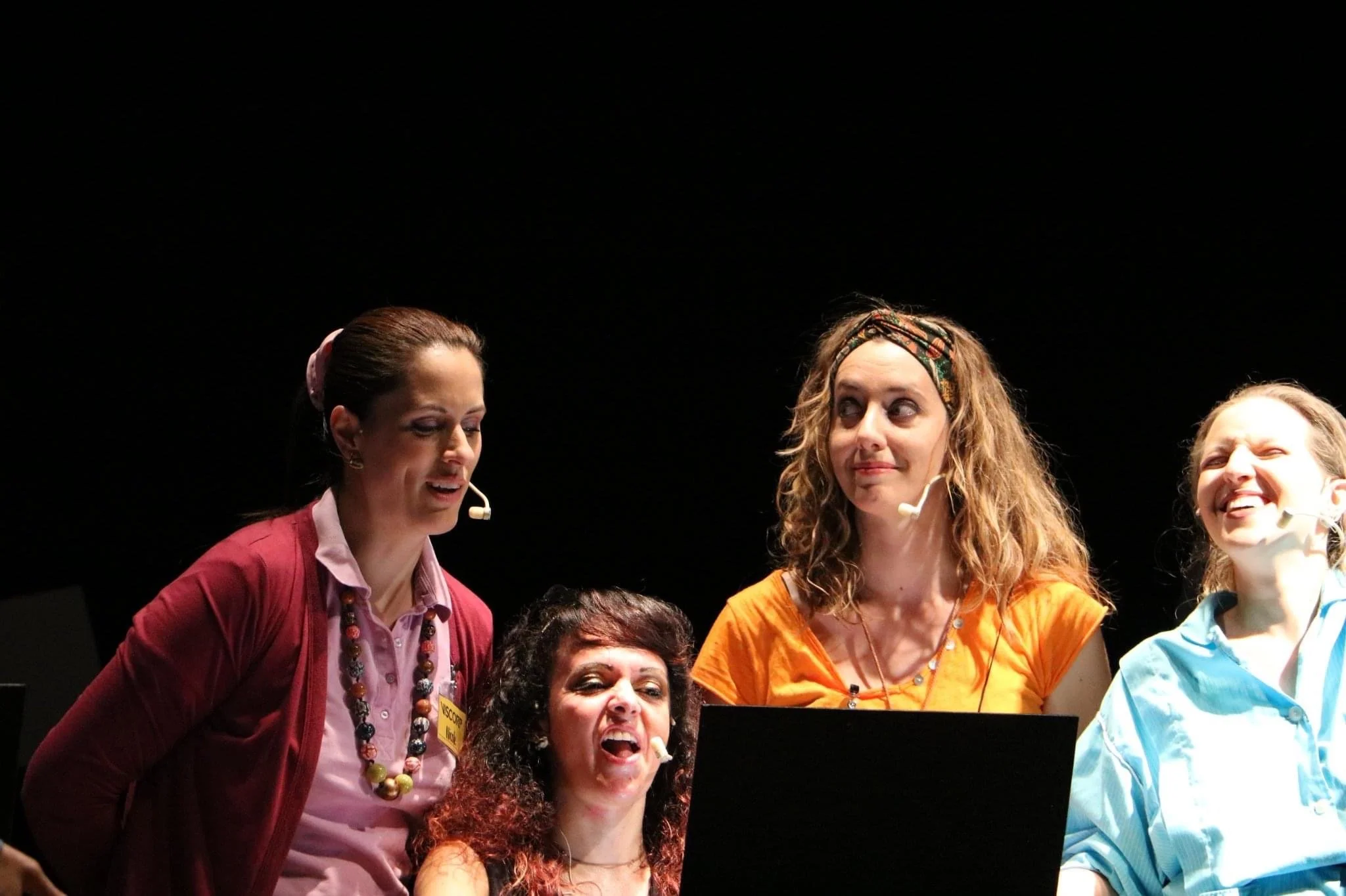 Group of women singing or performing on stage, smiling and engaged, wearing colorful clothing and accessories.