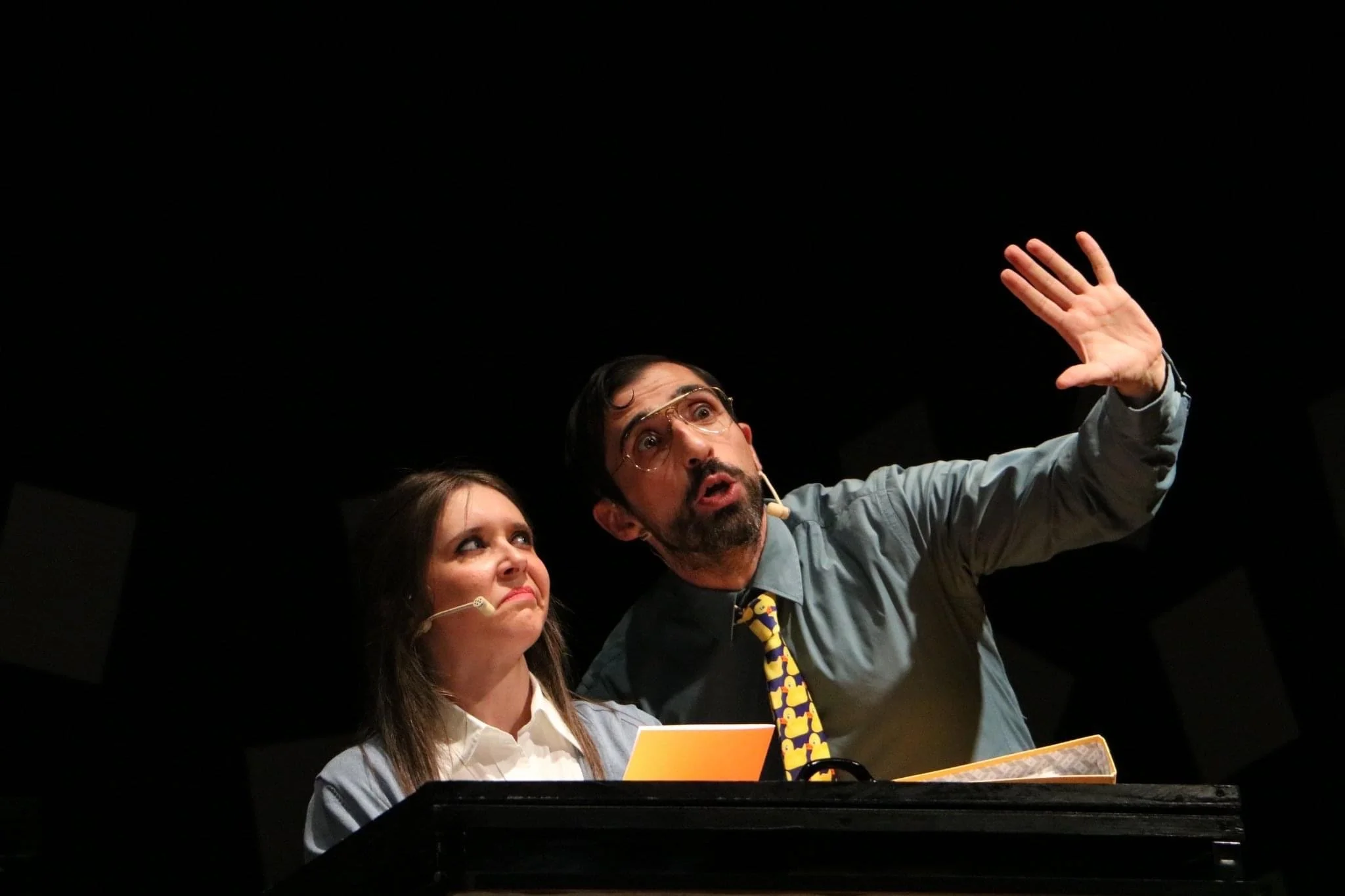 A man with glasses and a woman are on stage. The man is gesturing with his hand raised. The woman is looking at him, holding a yellow paper, both wearing microphones, with a black background.
