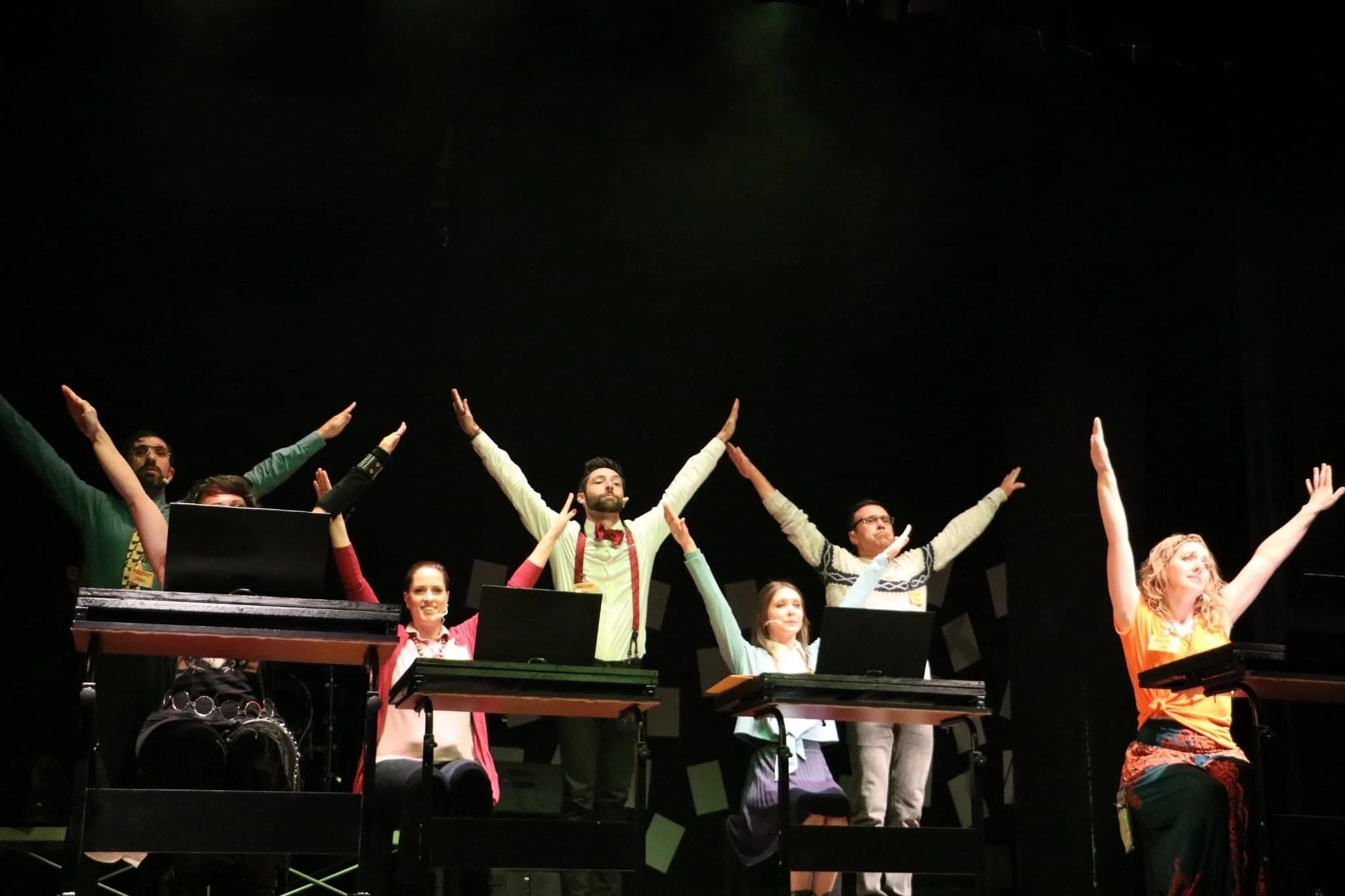 A group of people on stage with their arms raised during a performance, with a dark background and music stands or keyboards in front of them.