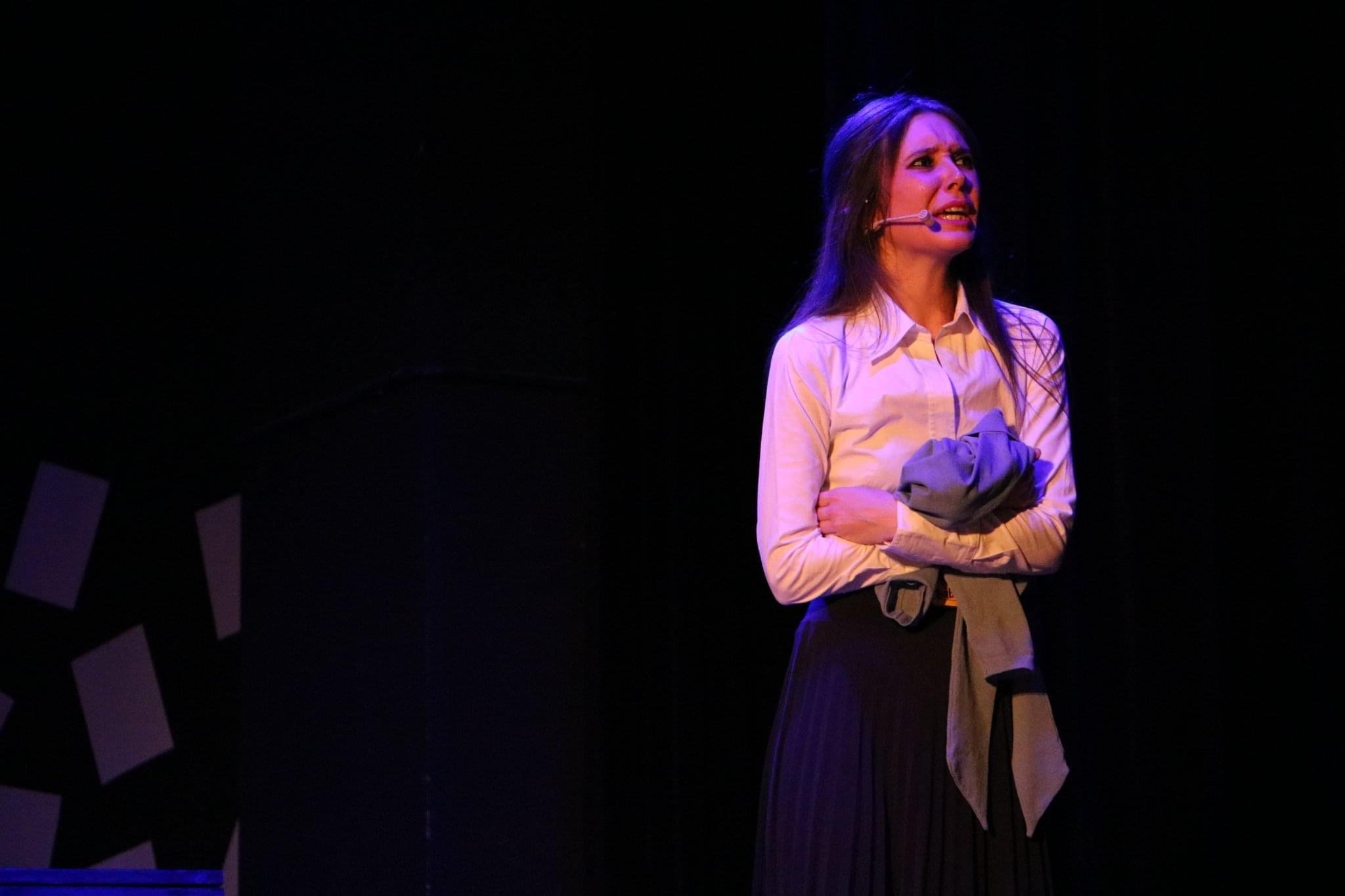 A woman wearing a white shirt and a long black skirt stands on stage with a microphone headset, appearing to perform or speak. She has long dark hair and is holding a gray jacket or piece of cloth in her crossed arms, with a dark background.