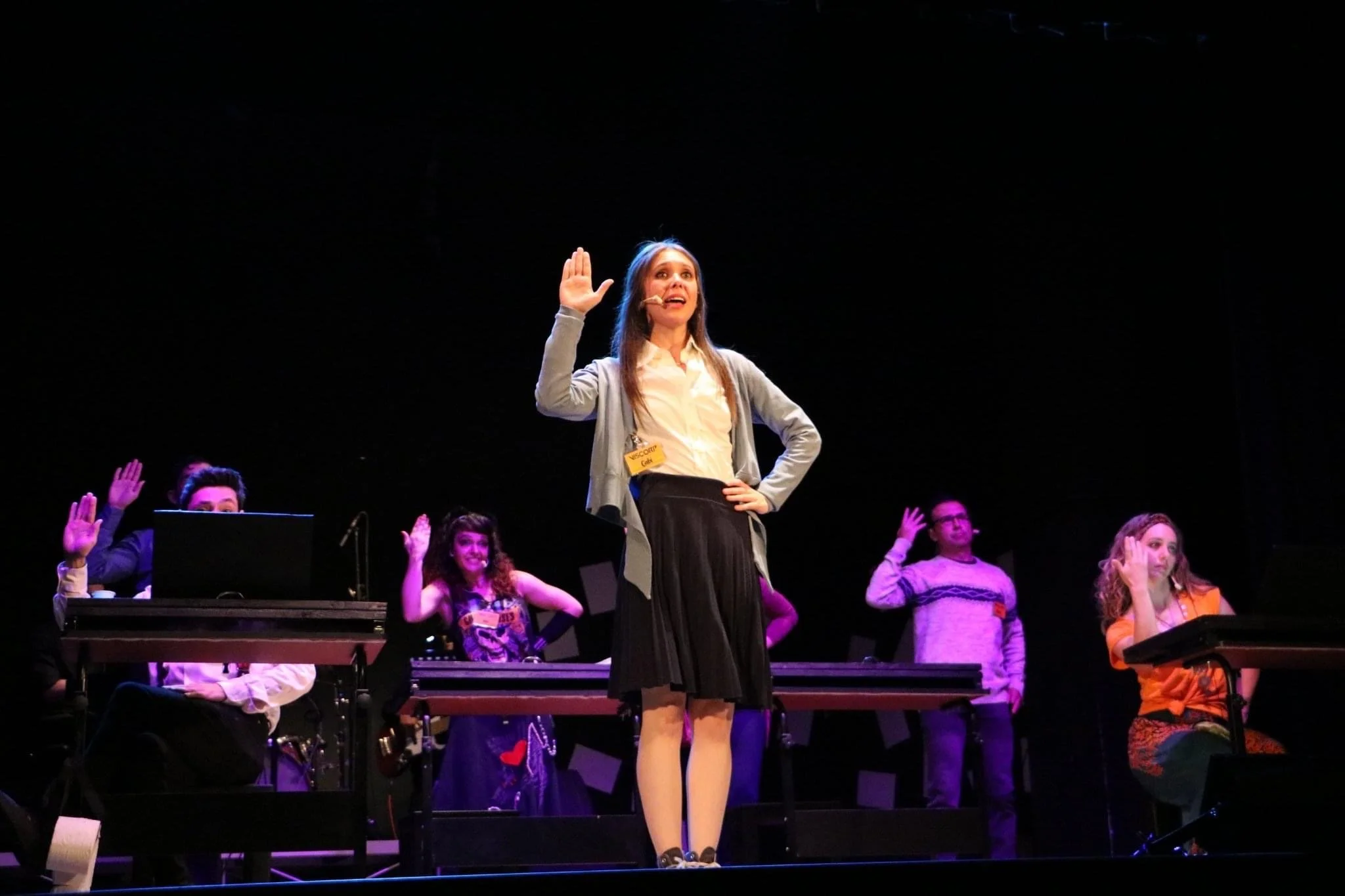 A woman with a microphone headset standing on stage with her right hand raised, singing or performing in front of an audience, surrounded by other performers.
