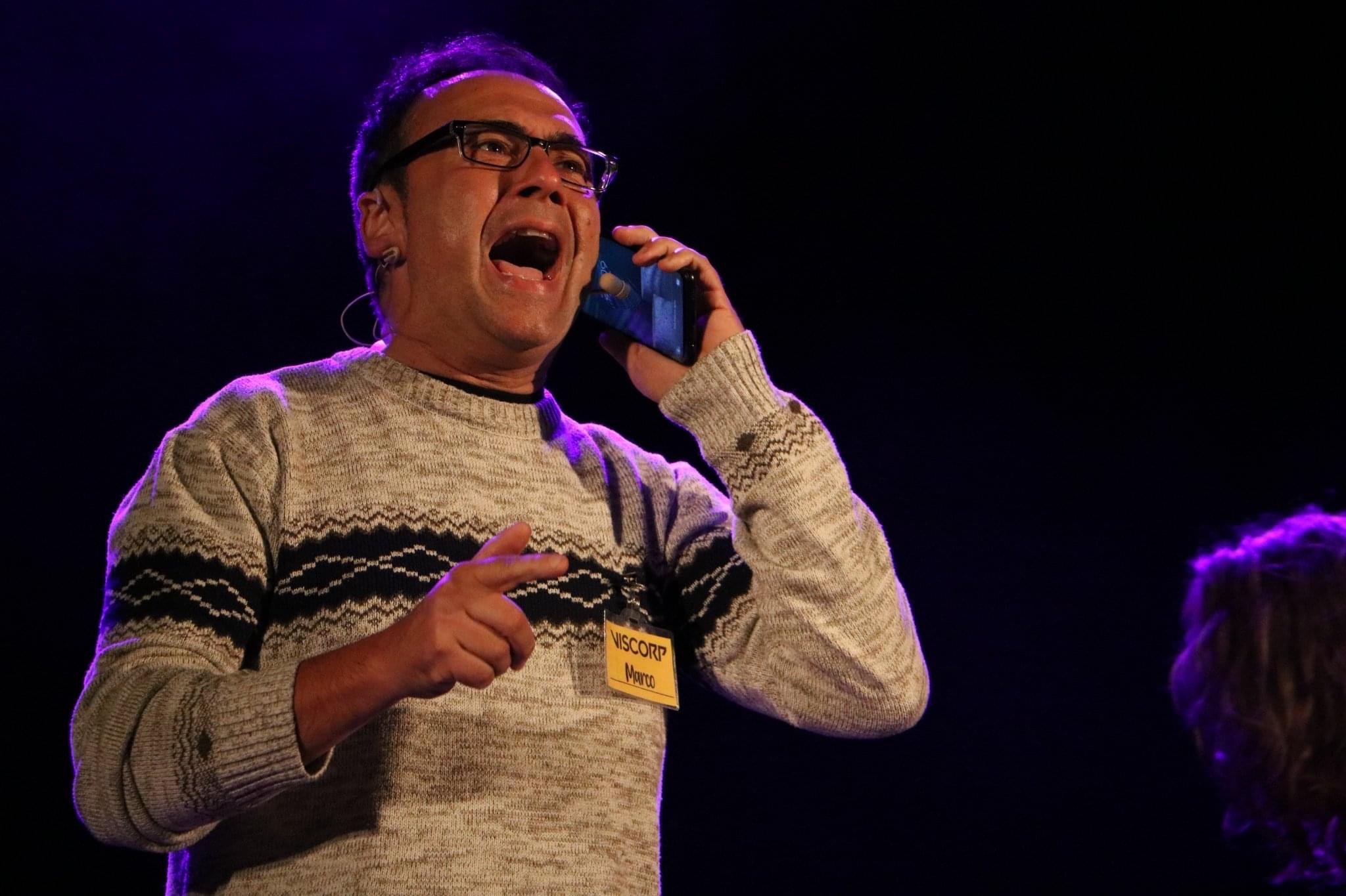 Man with glasses and a patterned sweater talking on a microphone during a conference.