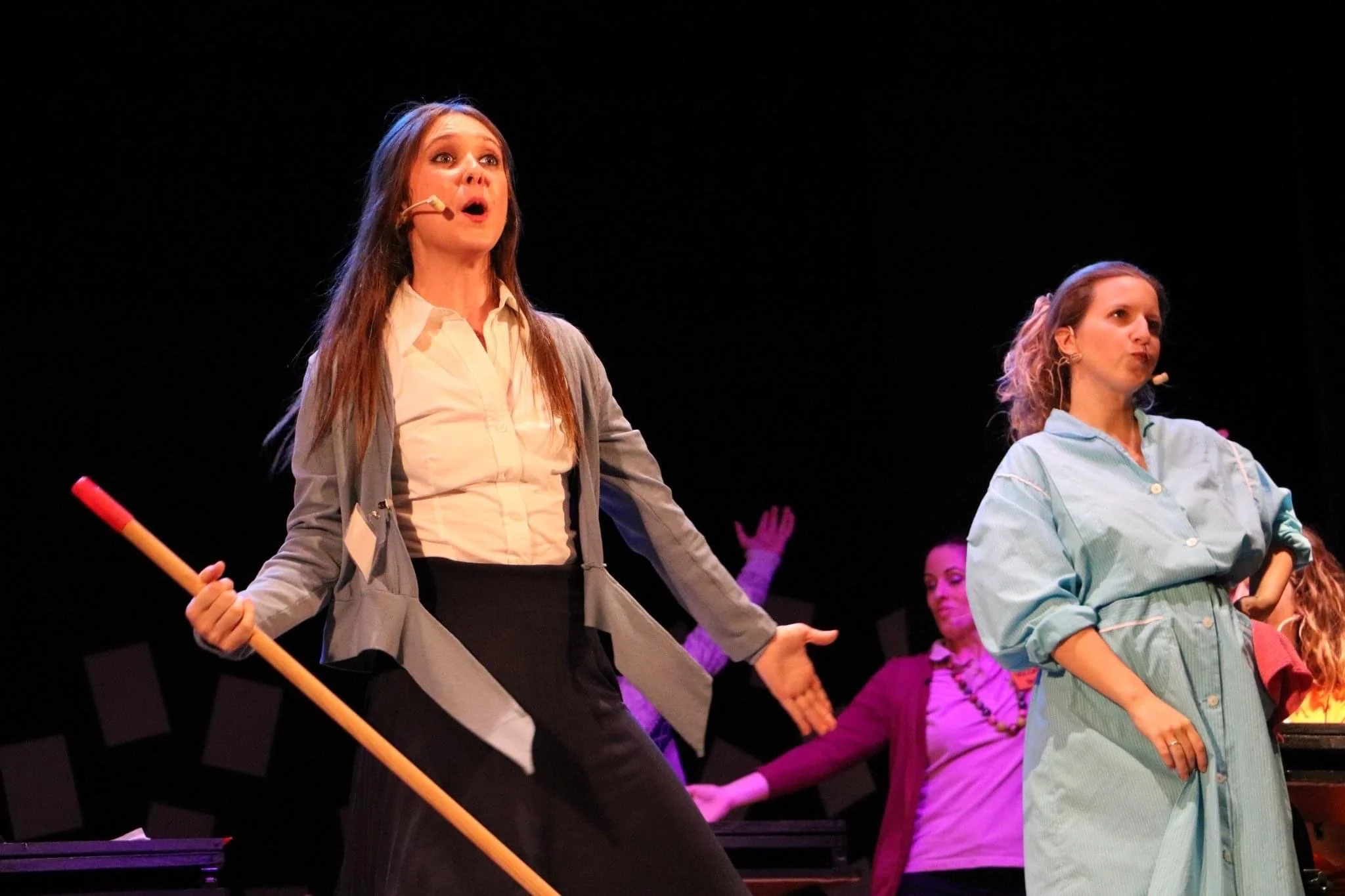 Women performing on stage, some holding sticks, under stage lighting with a dark background.
