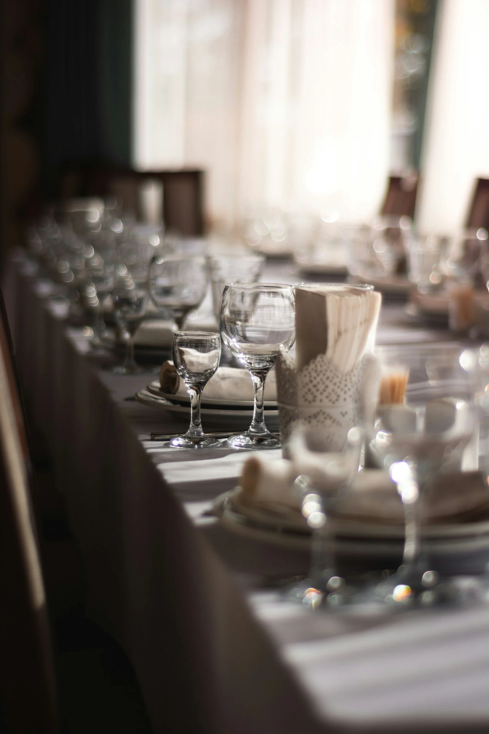 A table set with glassware, napkins, and tableware in a softly lit room with large windows and curtains, creating a warm, inviting atmosphere for dining.