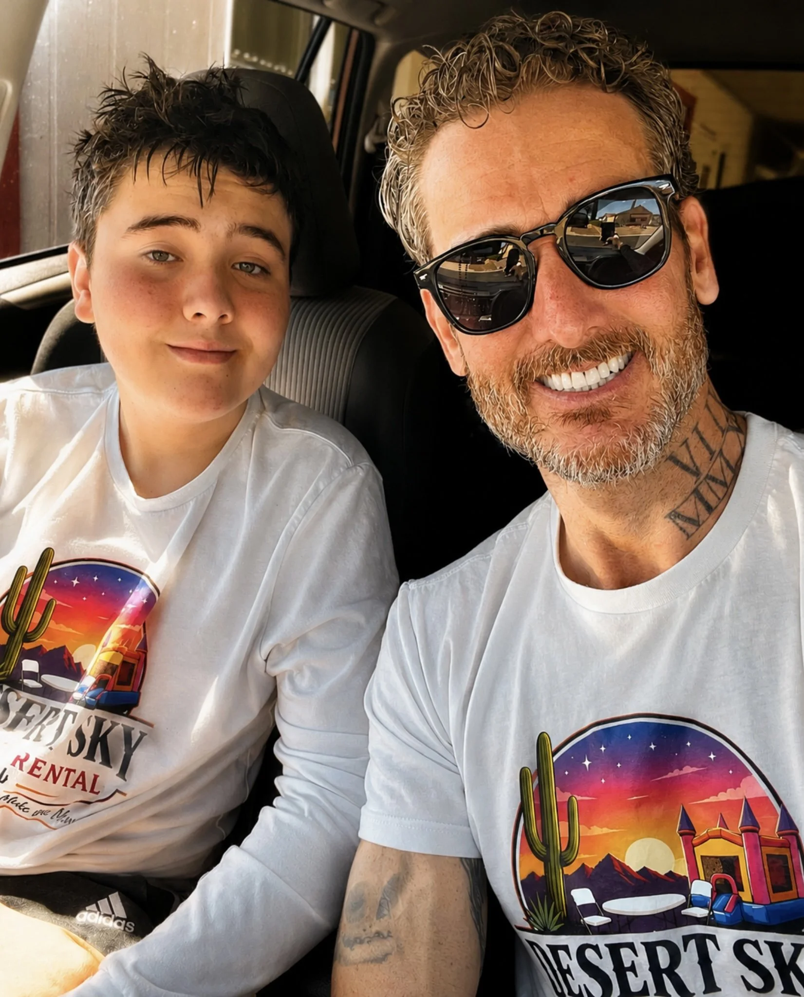 Two males taking a selfie inside a vehicle. The man on the right has curly hair, sunglasses, tattoos on his neck and arm, and is smiling. The boy on the left has short dark hair and is wearing a white shirt with a desert scene and colorful design.