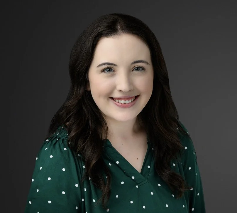 Young woman with dark brown hair smiling, wearing a green polka dot blouse, against a dark grey background.