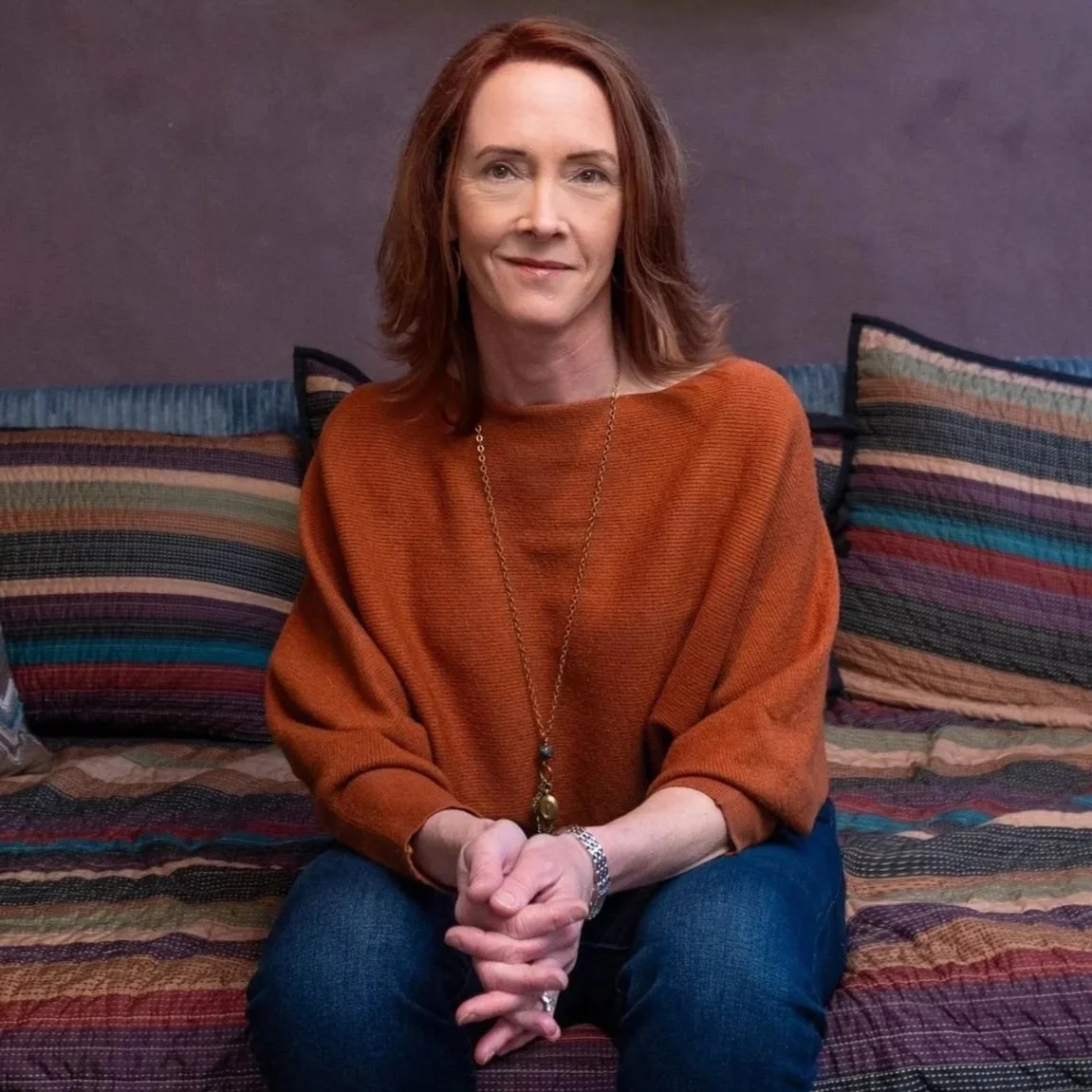 A woman with red hair sitting on a multicolored striped couch, smiling, wearing an orange sweater, jeans, a necklace, and a watch.