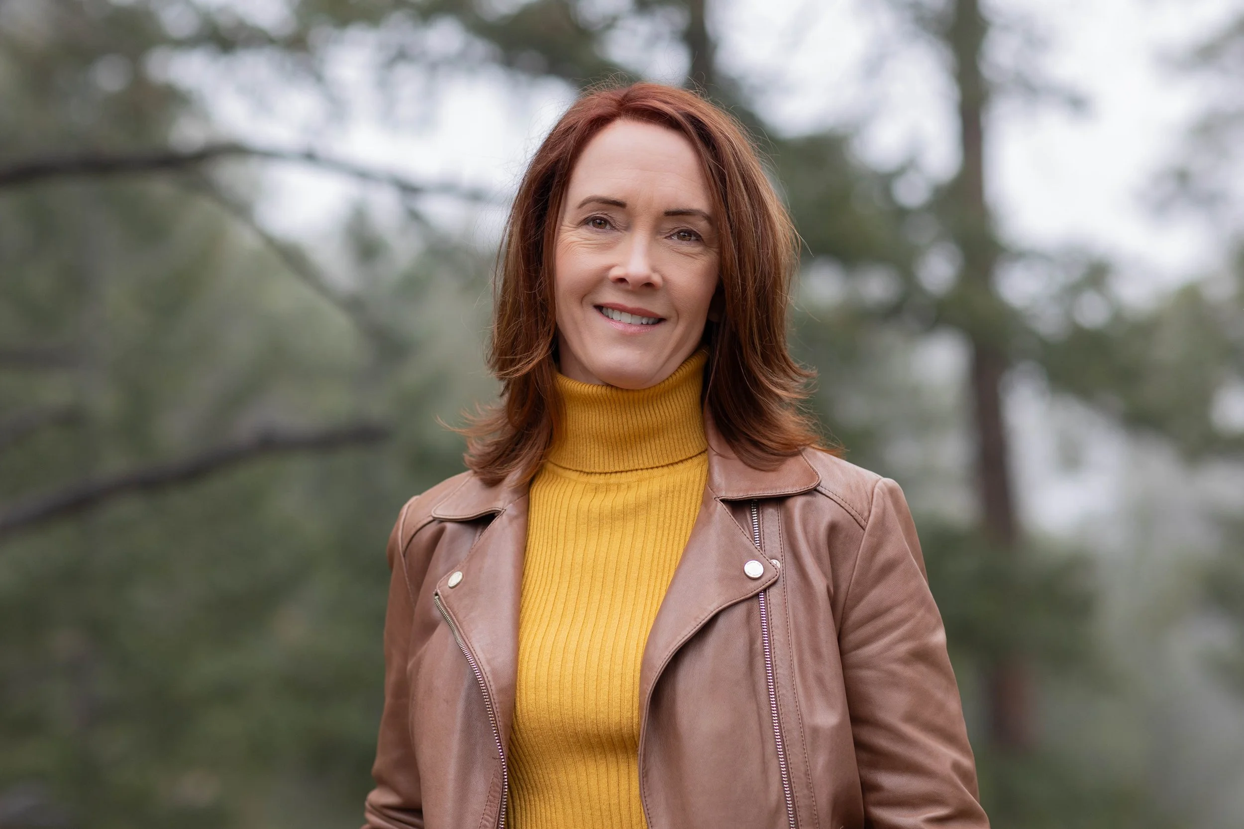 A woman with reddish-brown hair smiling outdoors, wearing a yellow turtleneck and a tan leather jacket, with a blurred background of trees.