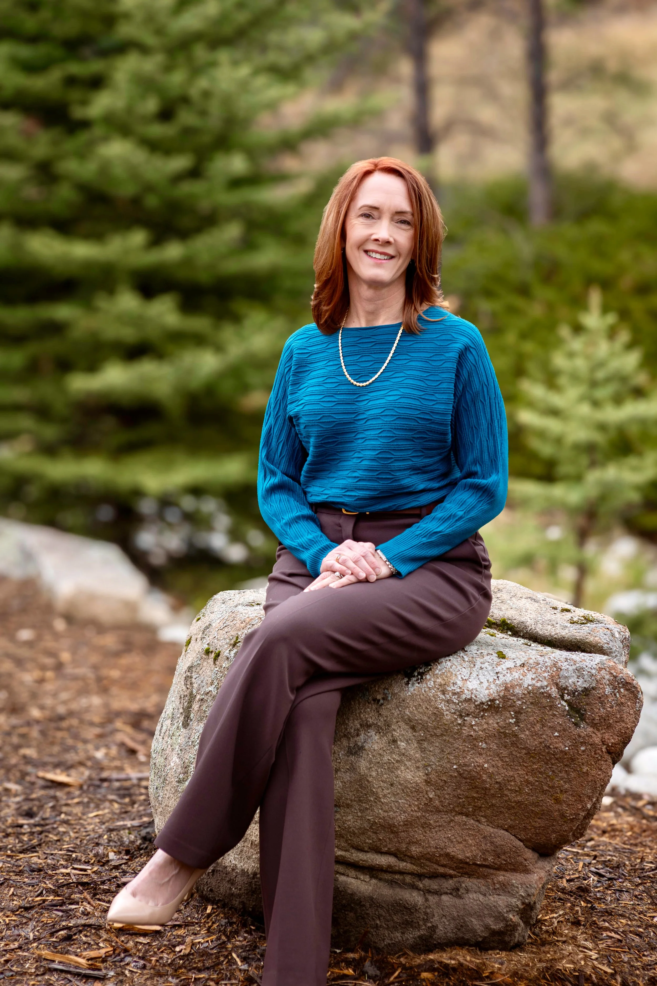 A woman with reddish hair wearing a blue top, dark brown pants, and beige heels, sitting on a large rock in a forested area, smiling at the camera.