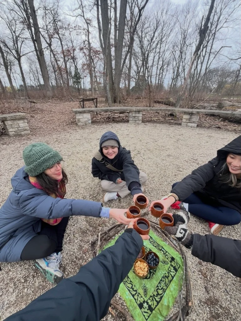 Four people in winter clothing gather outdoors around a blanket, toasting with wooden cups of tea beside snacks in a wooded park.