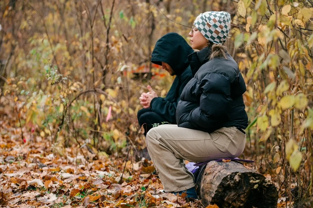 Two people in warm clothing sit on a log in an autumn forest.