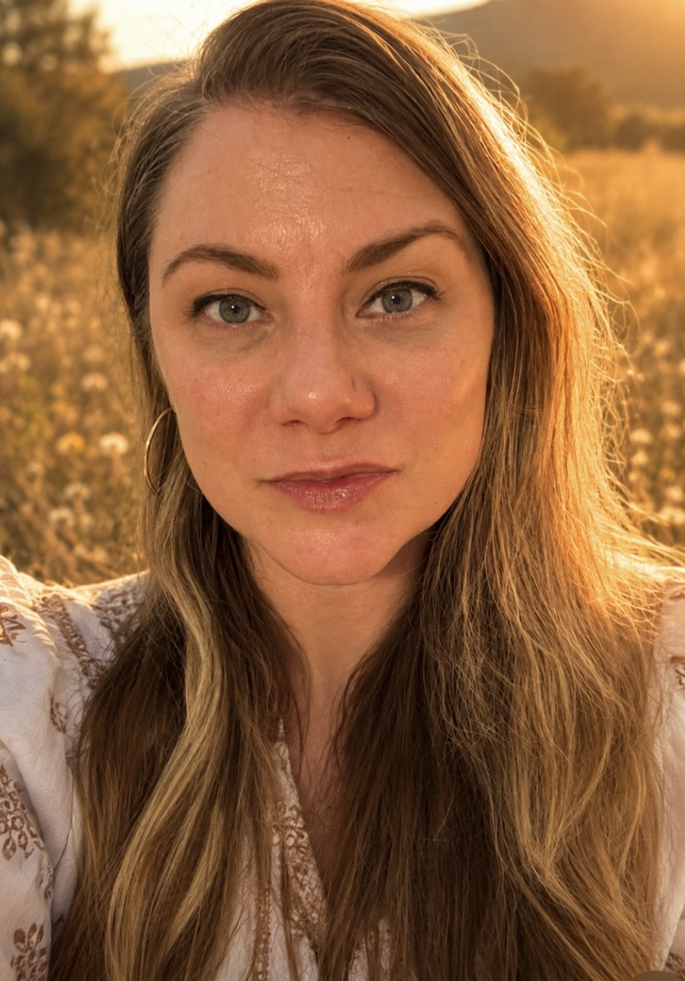 A woman with tan hair and blue eyes taking a selfie outdoors during sunset in a field of flowers, with mountains in the distance.