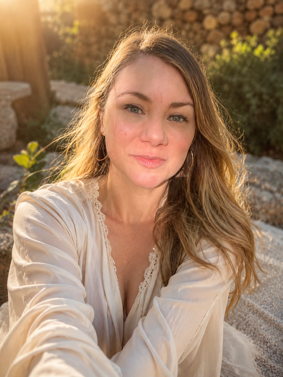 A woman with long, wavy hair and hoop earrings taking a selfie outdoors during sunset, wearing a cream-colored blouse