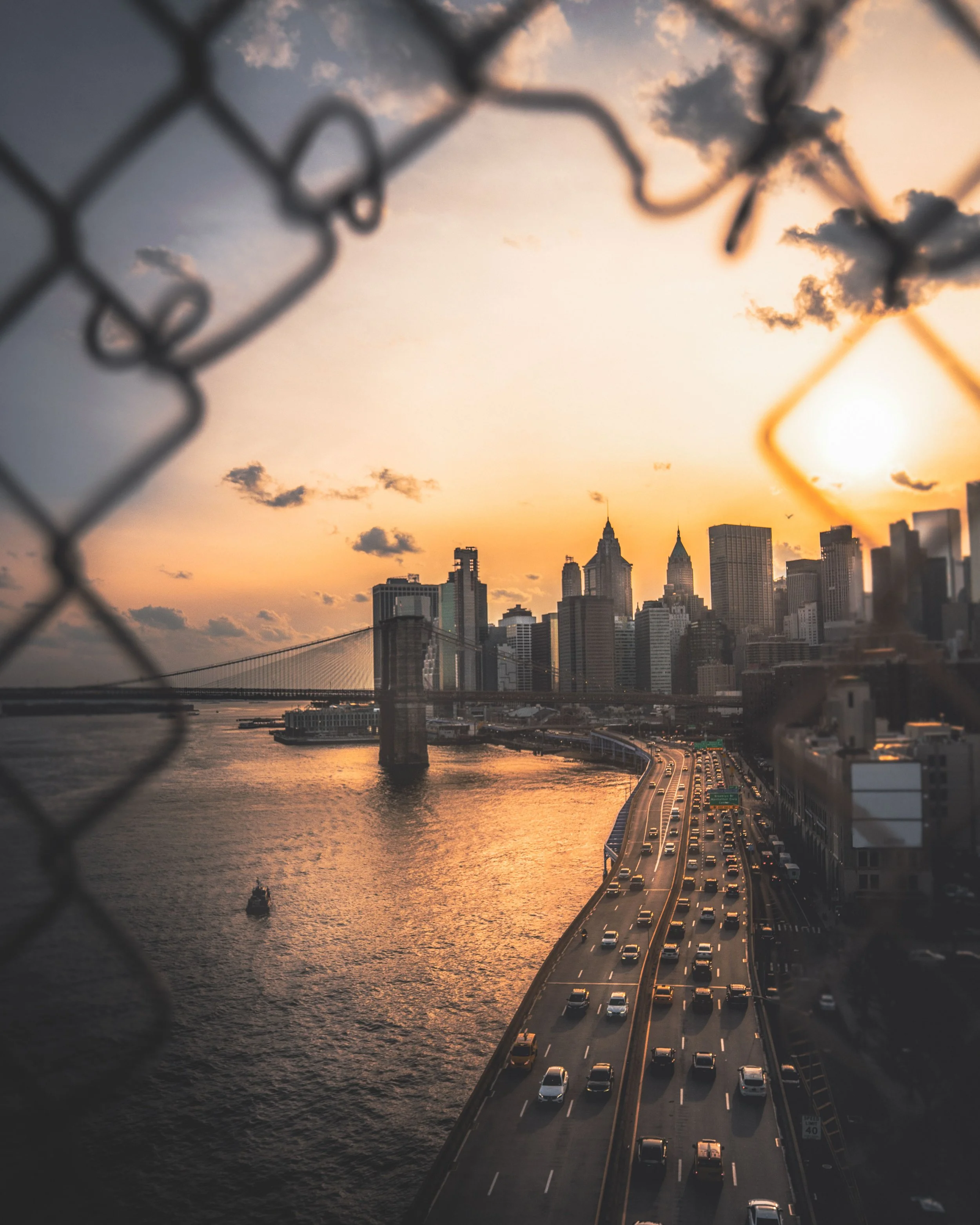 Photo of sunset over a city skyline, viewed through a chain-link fence. The city includes tall skyscrapers and a bridge over a river, with traffic on the bridge and warm sunlight illuminating the scene.