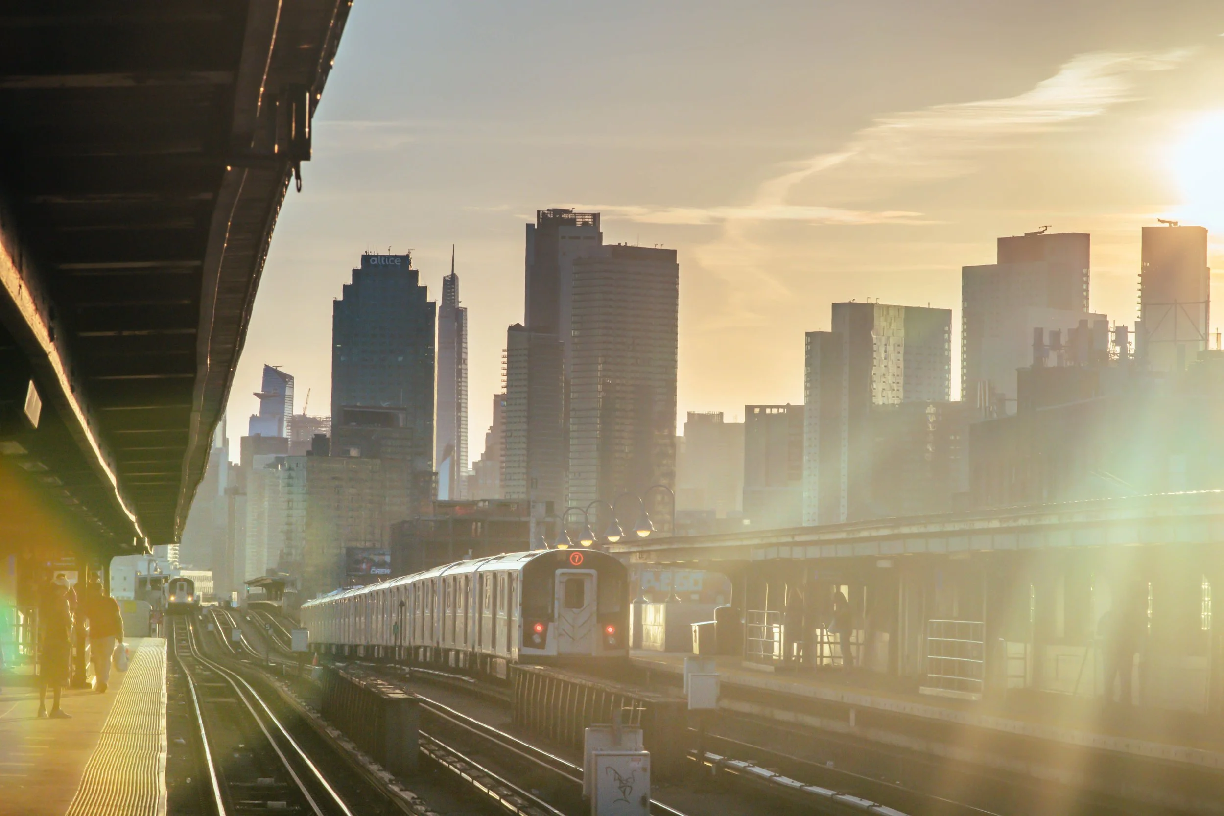 Urban scene at sunset showing skyscrapers, elevated train tracks, and a train approaching on the tracks with a city skyline in the background.