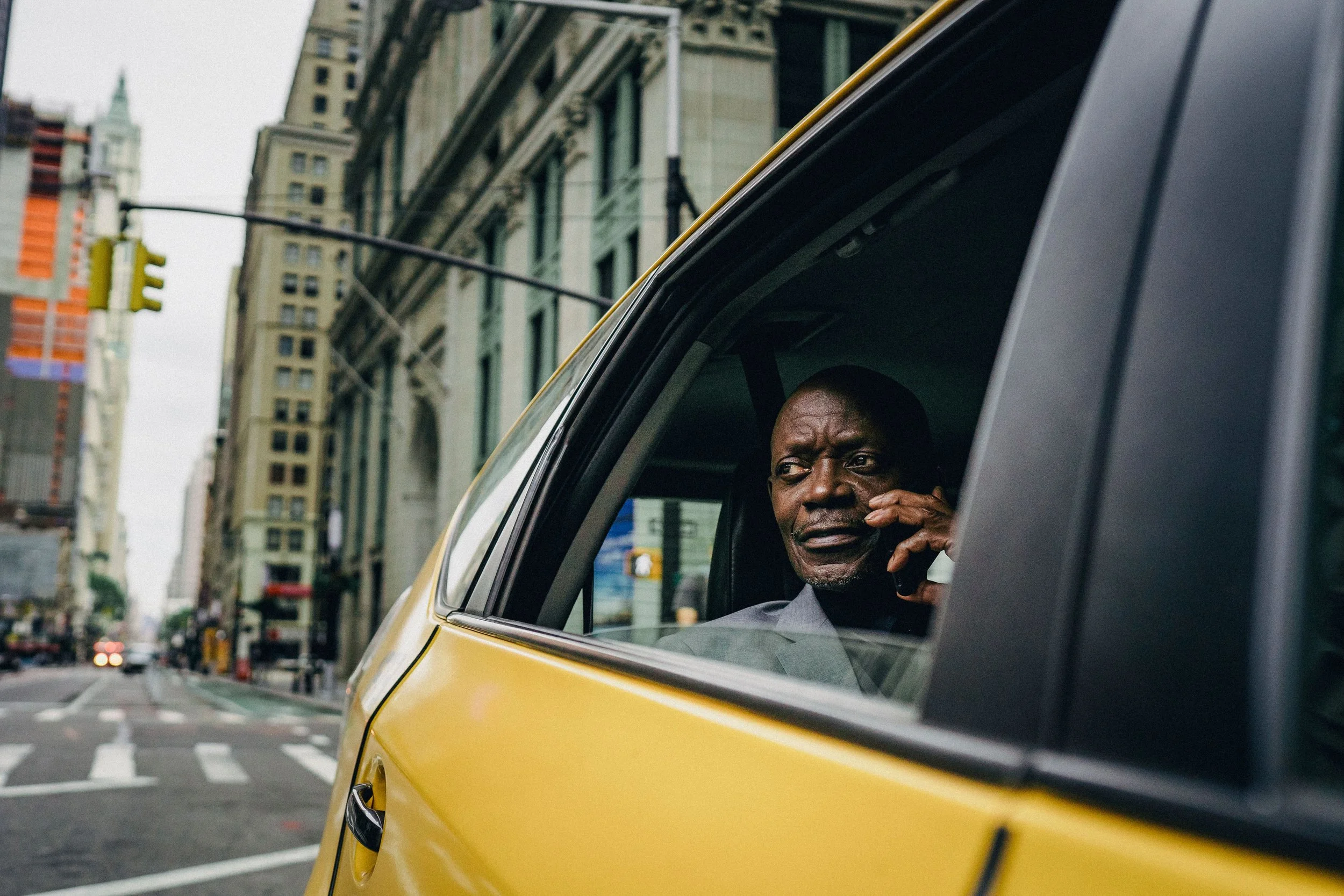 A man talking on a cellphone while sitting inside a yellow taxi cab, with city buildings and traffic lights in the background.