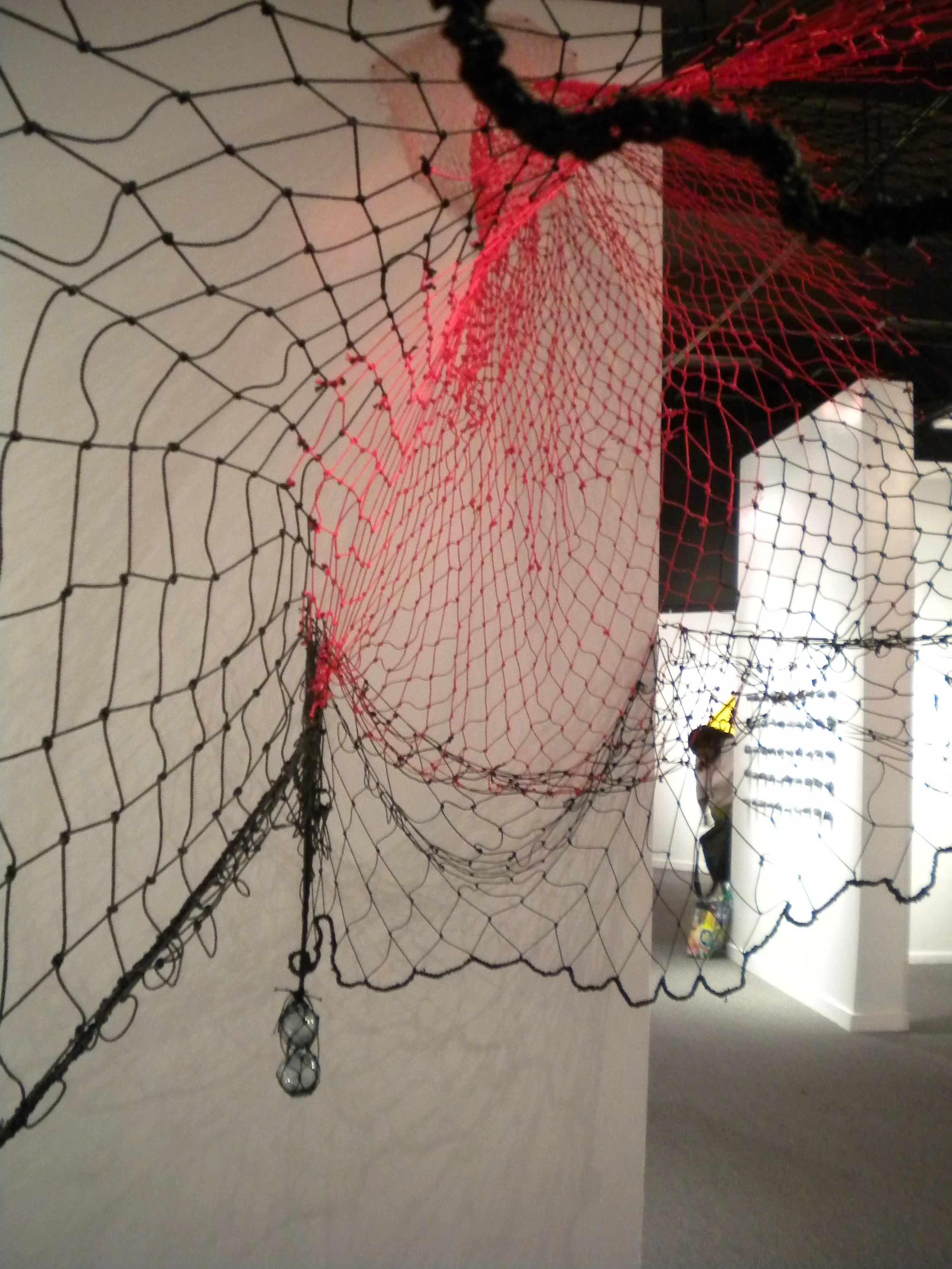 A decorative spider web with black, red, and blue strings, hanging in an indoor space with toys and furniture visible in the background.