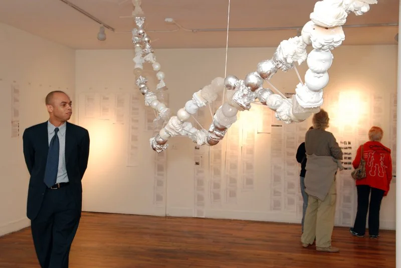 People viewing an art installation made of white and silver baby sculptures hanging from the ceiling  like a dna strand in an art gallery.