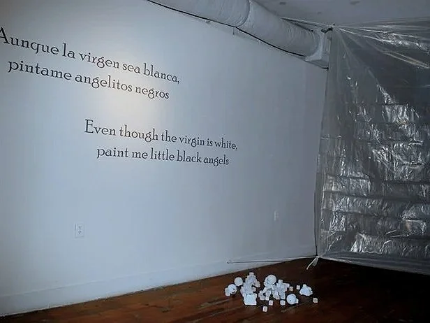 A white gallery wall with black text in Spanish and English, and a pile of small white broken dolls and blocks  on the wooden floor near a plastic sheet screen covering part of the gallery.